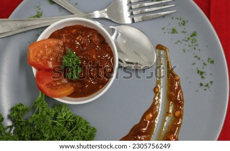 Indonesian style Smoked Tomato Sambal served in a white bowl garnished with parsley and tomato slices, on a gray melamine mat decorated with fresh parsley. Indonesian special ean chili sauce.