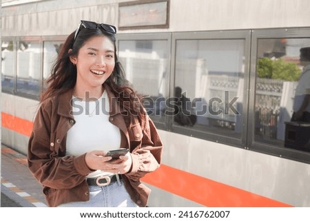 Indonesian young woman is smiling while holding her phone and standing in the station platform, next to a train