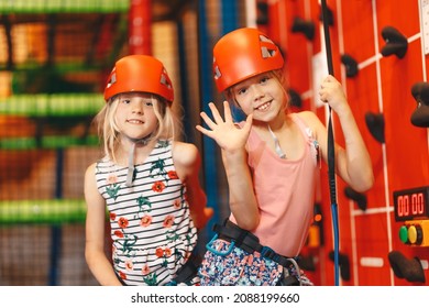 Indoor Climbing Class For Kids. School Girls Smiling At The Camera And Having Fun In Indoor Playground For Children. Two Happy Little Girls In Red Helmets Climbing The Wall In Bouldering Center