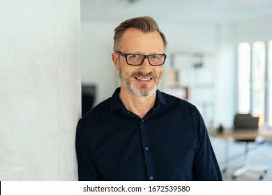 Indoor Close-up Portrait Of A Smiling Handsome Bearded Middle-aged Man In Glasses And Black Shirt, Leaning With His Shoulder On White Wall And Looking At Camera With Friendly Face, Standing In Office