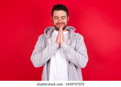 Indoor Closeup Of Young Handsome Caucasian Man In Sports Clothes Against Red Wall Practicing Yoga And Meditation, Holding Palms Together In Namaste, Looking Calm, Relaxed And Peaceful.