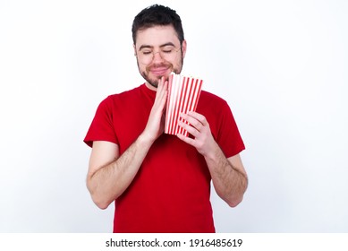 Indoor Closeup Of Young Handsome Man In Red T-shirt Against White Background Eating Popcorn Practicing Yoga And Meditation, Holding Palms Together In Namaste, Looking Calm, Relaxed And Peaceful.