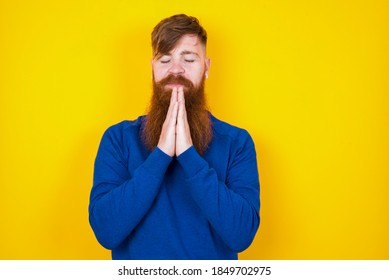 Indoor Closeup Of Young Handsome Red Haired Bearded Caucasian Man Standing Against Yellow Wall Practicing Yoga And Meditation, Holding Palms Together In Namaste Mudra, Looking Calm, Relaxed.