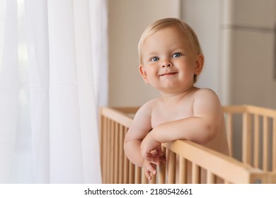 Indoor copy space portrait of adorable cute blue-eyed baby. Toddler standing in crib holding onto bumpers looking at his mom or dad with happy face and lovely smile.