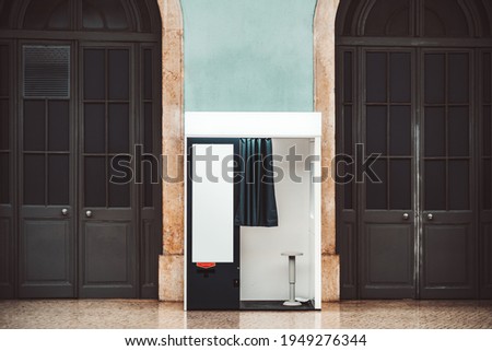 An indoor empty photo booth with open curtain, in between two wooden arch doors for creating photos for passport and other documents, with a blank template poster placeholder on the cabin wall