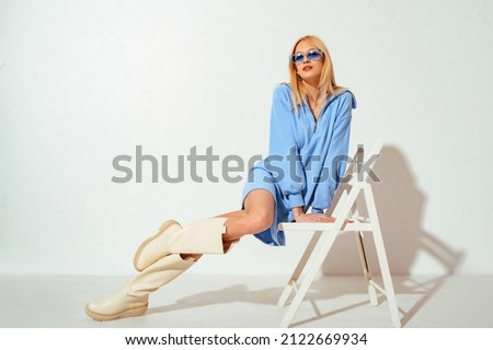 Indoor fashion portrait of beautiful blonde woman wearing trendy blue jersey mini dress with zip, color glasses, high leather boots, posing on white background. Copy, empty space for text