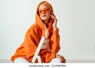 Indoor fashion portrait of young confident blonde woman wearing trendy orange hoodie, color sunglasses, posing on white background. Copy, empty space for text - Shutterstock ID 2119987892