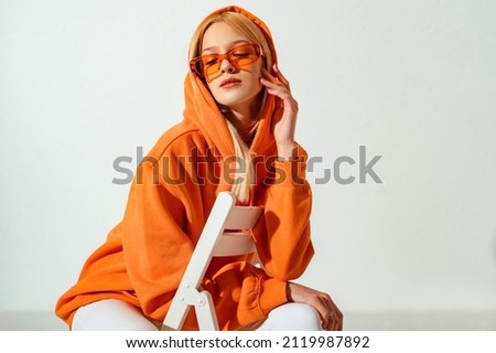 Indoor fashion portrait of young confident blonde woman wearing trendy orange hoodie, color sunglasses, posing on white background. Copy, empty space for text