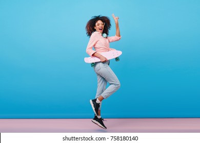 Indoor full-length portrait of confident african girl in pink shirt holding skateboard. Enthusiastic black woman with curly hairstyle posing in studio with blue interior.