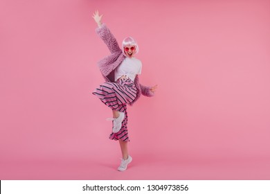 Indoor full-length portrait of pleasant glamorous girl with pink hair. Happy young lady in periwig dancing in white sneakers in studio and laughing.