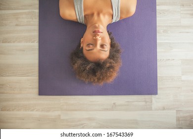 Indoor High-angle Shot Of An African Woman Lying On A Yoga Mat With Her Eyes Closed.