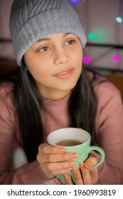 Indoor Image Of An Asian, Indian Beautiful, Serene Young Woman Looking Away And Contemplating While Drinking And Holding A Cup Of Coffee In Winter Season At Home. She Is Wearing Warm Clothes Sweater.