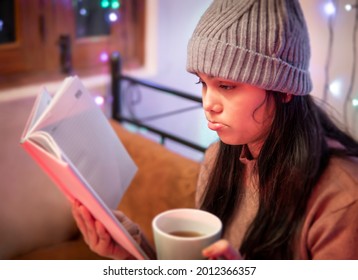 Indoor Image Of An Asian, Indian Beautiful Unhappy Young Woman Reading A Book While Drinking Coffee In Winter Season At Home. She Is Wearing Warm Clothes, Sweater And A Woolen Cap.