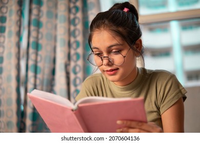 Indoor Image Of An Asian, Indian Happy Young Woman With Round Eyeglasses Sitting In Her Room And Reading A Book. She Is Studying Books And Doing Preparation For The Exam.