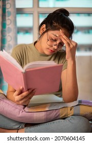 Indoor Image Of Asian, Indian Young Woman With Round Eyeglasses Sitting In Her Room And  Having Bad Headache While Reading A Book And Holding Her Head Out Of Headache During Preparation For The Exam