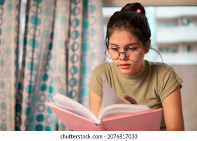 Indoor Image Of An Asian, Indian Young Woman With Round Eyeglasses Sitting In Her Room And Reading A Book. She Is Studying Books And Doing Preparation For The Exam.