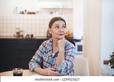 Indoor image of attractive serious mature sixty year old housewife wearing casual checkered shirt having coffee, sitting at kitchen table with mug and looking sideways with thoughtful expression