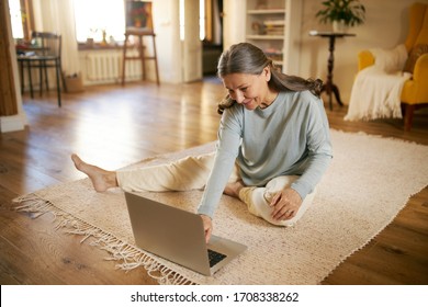 Indoor image of beautiful energetic female on retirement sitting barefoot on floor using laptop turning on calm music for meditation. Elderly European woman surfing internet on portable computer