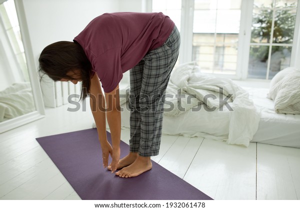 Indoor\
image of brunette dark skinned female standing barefooted on yoga\
mat stretching body in the morning posing in cozy bedroom, bending\
forwards, trying to touch toes with her\
fingers