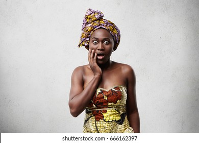 Indoor image of dark-skinned African woman wearing traditional clothes having stupefaction look. Surprised black woman looking with bugged eyes and opened mouth holding hand on cheek isolated on white