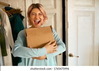 Indoor image of happy cheerful young woman holding cardboard box delivered to her apartment, expressing excitement, going to unpack parcel, having impatient overjoyed look. Food delivery and shopping