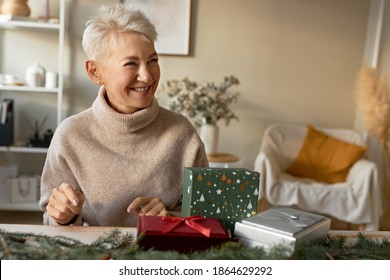 Indoor image of happy gray haired female pensioner laughing being in good festive mood, enjoying Christmas preparations, sitting at table with boxes, fir branches, wrapping presents for family