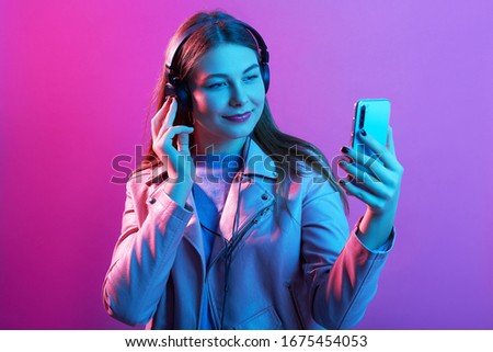 Indoor image of pleasant positive young female taking picture for social networking sites, having headphones, posing isolated over neon background, wearing casual clothes. Technology concept.