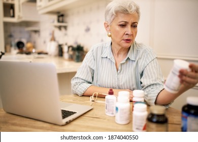 Indoor Image Of Short Haired Serious Senior Female Sitting At Table With Laptop And Health Supplements, Reading Nutrition Label On Bottle, Taking Vitamins Before Meal. Technology, Health And Diet