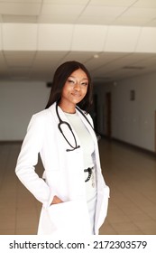 Indoor Image Of Young Black African Female Medical Professional Wearing White Coat Uniform And Holding Stethoscope Standing In Clinic Hall. Concept Of Racial Bias And Underserved Communities Society