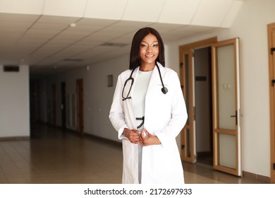 Indoor Image Of Young Black African Female Medical Professional Wearing White Coat Uniform And Holding Stethoscope Standing In Clinic Hall. Concept Of Underrepresented Minorities Racial Disparities