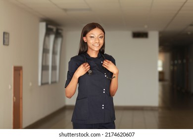 Indoor image of young black African female medical professional wearing dark blue uniform and holding stethoscope standing in clinic hall. Concept of underrepresented minorities racial disparities