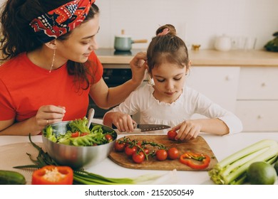 Indoor image of young good-looking brunette mother in red bandana and t-shirt teaching her daughter in white blouse to cook, touching her hair gently while she slicing cherry tomato for salad - Powered by Shutterstock