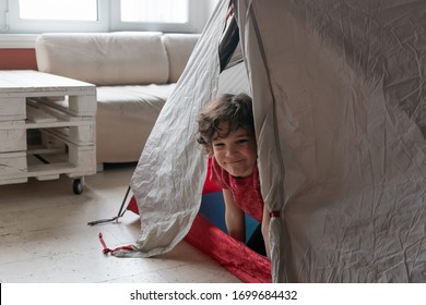 Indoor Life At The Coronavirus Quarantine. Cute Curly Boy Looks From A Tent At Home.