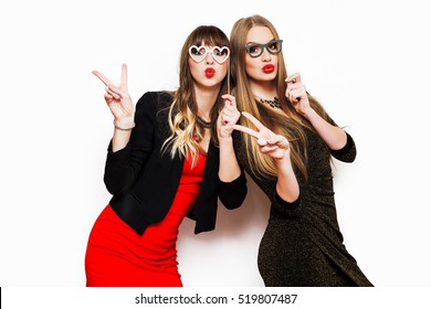  Indoor  lifestyle portrait of two bed friends, elegant women in evening dress Holiday makeup and bright party accessories posing on white wall, enjoying birthday or new year party. 