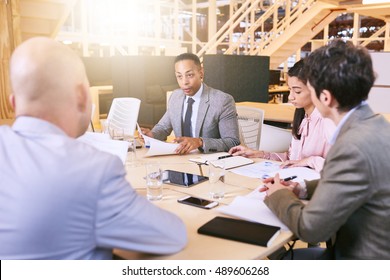 Indoor Meeting Between 4 Business Executives In A Warm Creative Environment, Making Use Of Tech And Documents During The Early Hours Of The Morning In Preparation For The Coming Week.