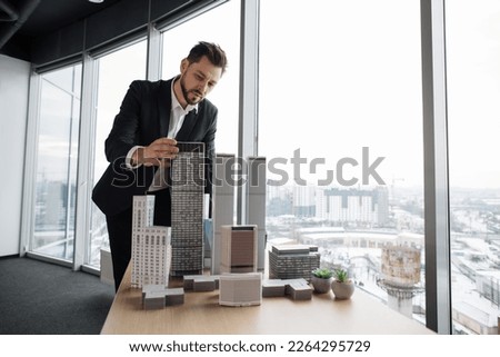 Indoor office portrait of bearded young business man, wearing white shirt and black suit, standing near wooden table with 3D megalopolis model in modern office interior with panoramic city view. [[stock_photo]] © 
