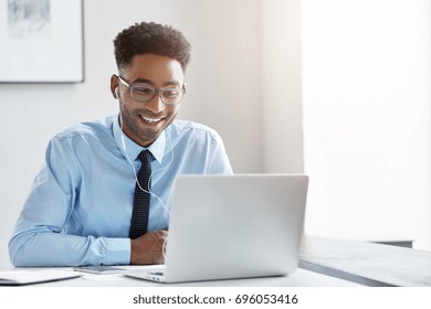 Indoor office shot of cheerful successful young African American manager with stubble sitting in front of open laptop wearing earphones while having video conference call with business partners - Powered by Shutterstock