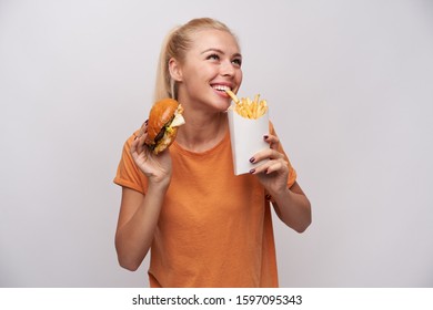 Indoor photo of attractive young blonde lady in orange t-shirt smiling cheerfully while pulling with teeth french fries and keeping hamburger in other hand, isolated over white background