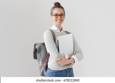 Indoor Photo Of College Student Girl Isolated On Gray Background, Smiling At Camera, Pressing Laptop To Chest, Wearing Backpack, Ready To Go To Studies, Start New Project And Suggest New Ideas.