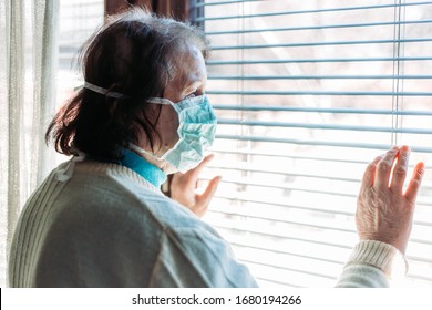 Indoor Photo Of Elderly Woman With Medical, Surgical Mask On Her Face Looking Through The Window Outside, Looking Worried. Quarantine Time