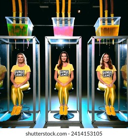 Indoor photo of group of 3 caucasian women aged 25 wearing tight yellow t shirts and yellow lycra shorts and their t shirts have "gunge games" written on them. they are on a dungeon style gameshow. they are sitting inside three tall glass cubicles with
