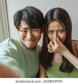 Indoor photo of medium shot from phone, east asian young couple with pastel green clothes having a selfie at home, the guy is smiling obliviously holding the phone, the girl is grimacing and holding her nose due to his bad body odor, eye level shot,