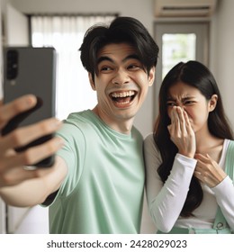 Indoor photo of medium wide shot from phone, east asian young couple with pastel green clothes having a selfie at home, the guy is smiling obliviously holding the phone, the girl is grimacing and covering her nose due to his bad body odor, eye level shot,