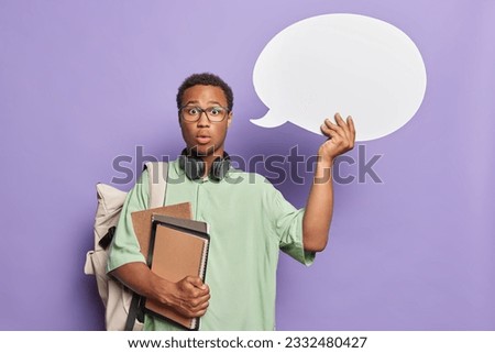 Indoor photo of young African male student with books notebooks tablet standing in centre isolated on purple background holding white speechbubble surprised to get test results. People emotions