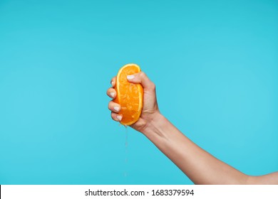 Indoor photo of young attractive hand holding orange and clenching a fists while queezing juice, preparing breakfast while posing over blue background