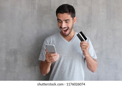 Indoor photo of young European Caucasian man isolated on grey background holding smartphone and plastic card in his hands looking at screen with amazement, happy because ecommerce is so easy