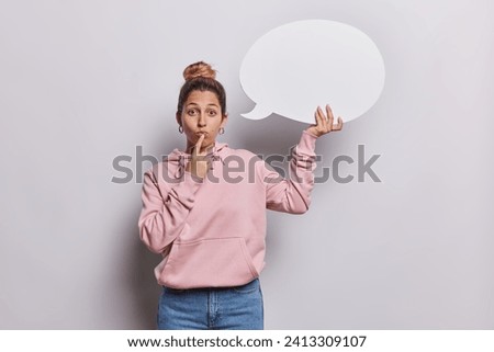 Indoor photo of young pretty European girl standing in centre on white background wearing pink hoodie and blue jeans holding speech bubble with space for promotion expressing positive emotions