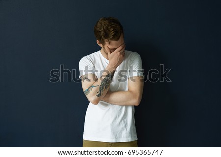 Indoor picture of attractive shy embarrassed young man with beard and tattoo on right arm making faceplam gesture, feeling shamed about something. Human reaction, feelings and attitude concept