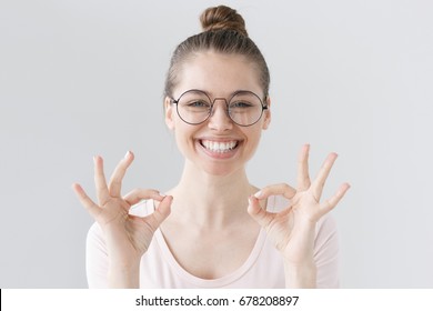 Indoor picture of beautiful girl isolated on gray background wearing big round spectacles, looking happy and positive as she is showing double ok sign, being sure about best outcomes in future. - Shutterstock ID 678208897