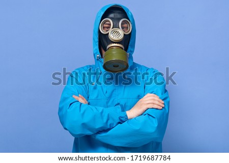 Indoor picture of brave guy standing isolated over blue background in studio, posing with folded arms, wearing gas mask and protection suit, being on secret mission. Chernobyl disaster concept.